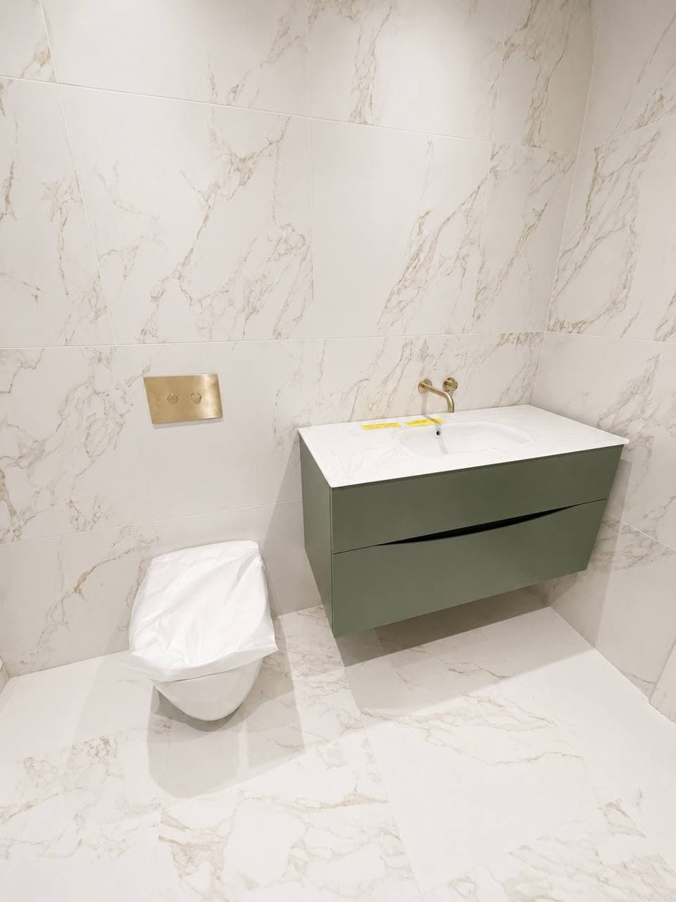 Wall-mounted green vanity and toilet surrounded by marble-effect tiling and brushed brass fittings